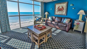 A room with a large window overlooking a beach, a brown leather sofa, a wooden coffee table, and a patterned carpet. - ⋆𖦹NEW❀Amazing Direct OceanFront/Spacious/Pristine/Indr-WaterParks 2BR2BA2Balcs (Myrtle Beach)