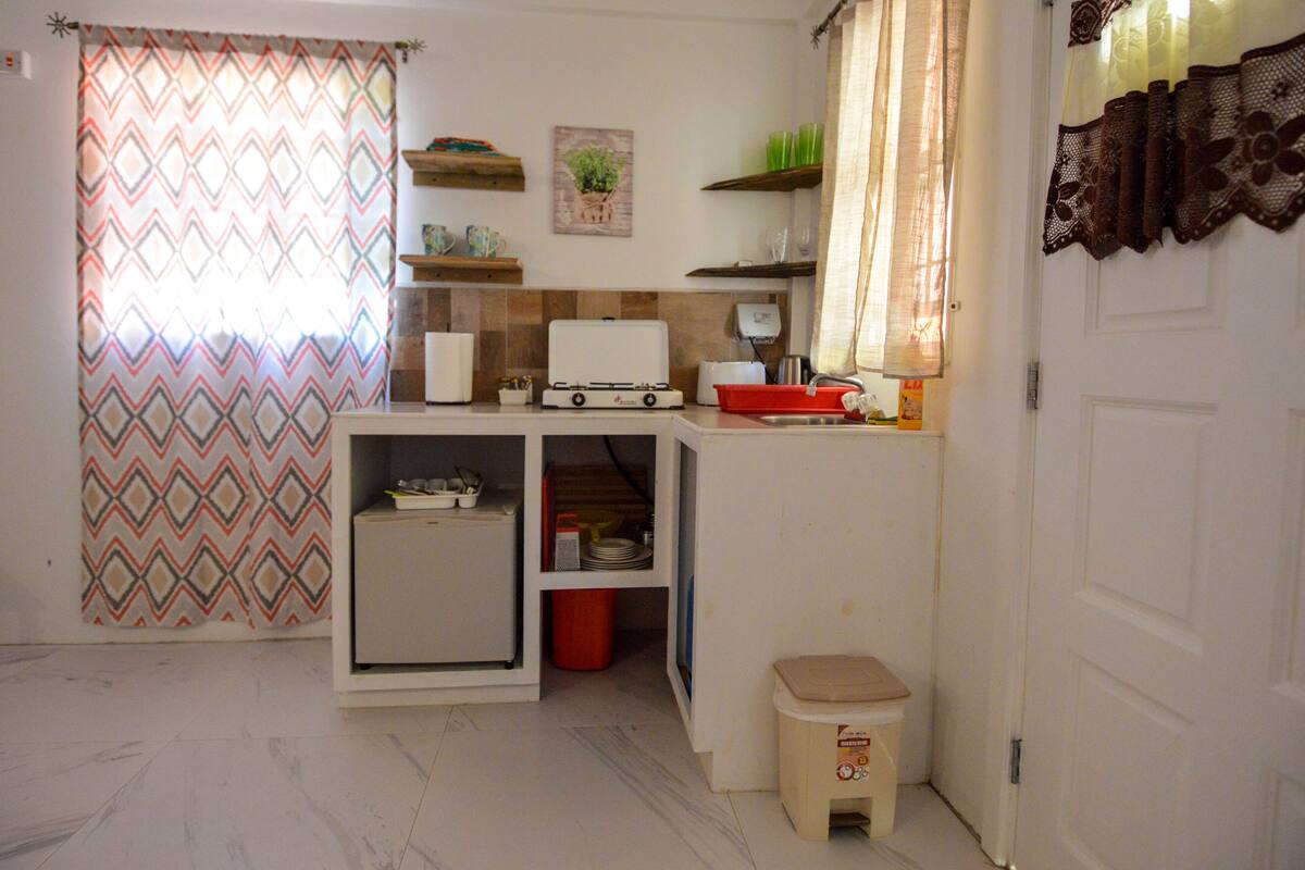 Une cuisine compacte équipée d’un réfrigérateur, d’un four à micro-ondes et d’un évier. Des étagères se trouvent au-dessus du plan de travail et un petit tabouret est placé à côté de la porte.