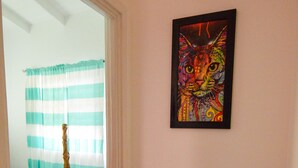 Um corredor com uma porta de madeira que dá para um quarto com cortina listrada. Um quadro colorido de um gato está pendurado na parede, e um vaso alto está colocado no chão.
