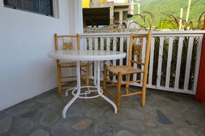 A small outdoor seating area with a round table and two wicker chairs, set against a white fence with a view of a hillside. - Frenz (Soufrière)