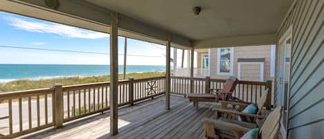 A wooden deck with a view of the ocean, a covered porch with chairs, and a building with a window.
