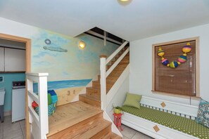 Escadaria que leva a um quarto com mural temático praiano, uma janela com decorações coloridas e um pequeno espaço de estar com um almofada listrada.