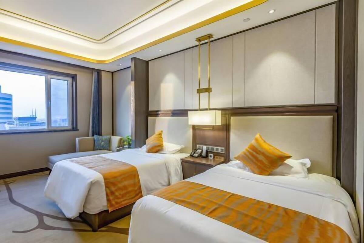 A hotel room with two beds, a nightstand, a lamp, and a view of the city.
