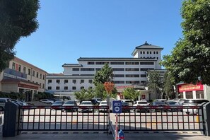 Un edificio de varios pisos con entrada protegida por una reja, rodeado de coches aparcados y árboles.