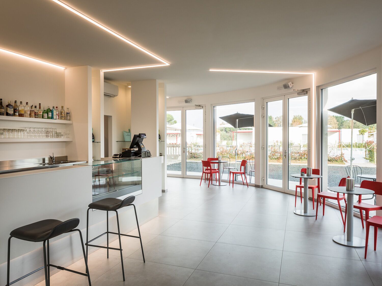 A modern bar area with a counter, stools, and a coffee machine. There are red chairs and tables outside, with a view of the garden.