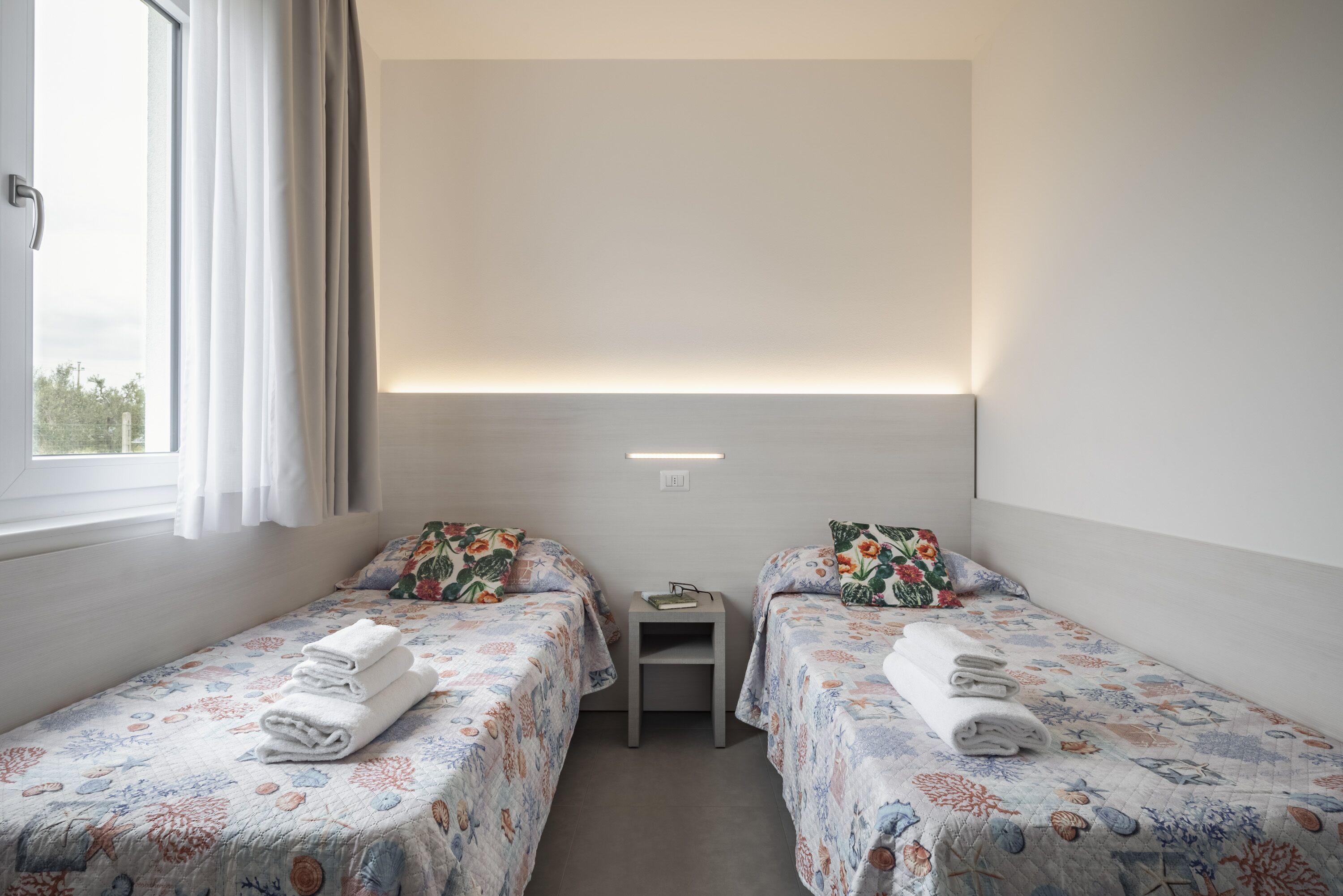 Una habitación con dos camas, cada una con ropa de cama y toallas estampadas con motivos florales. Hay una mesita entre las camas y una ventana a la izquierda.