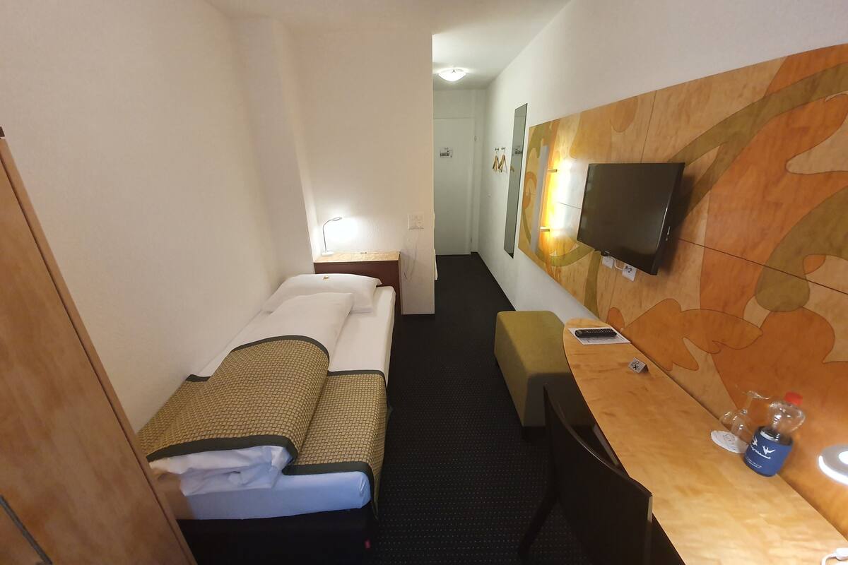 Ein Hotelzimmer mit Bett, Schreibtisch mit Stuhl, einem an der Wand befestigten Fernseher und einem kleinen Tisch mit Flasche und Fernbedienung.