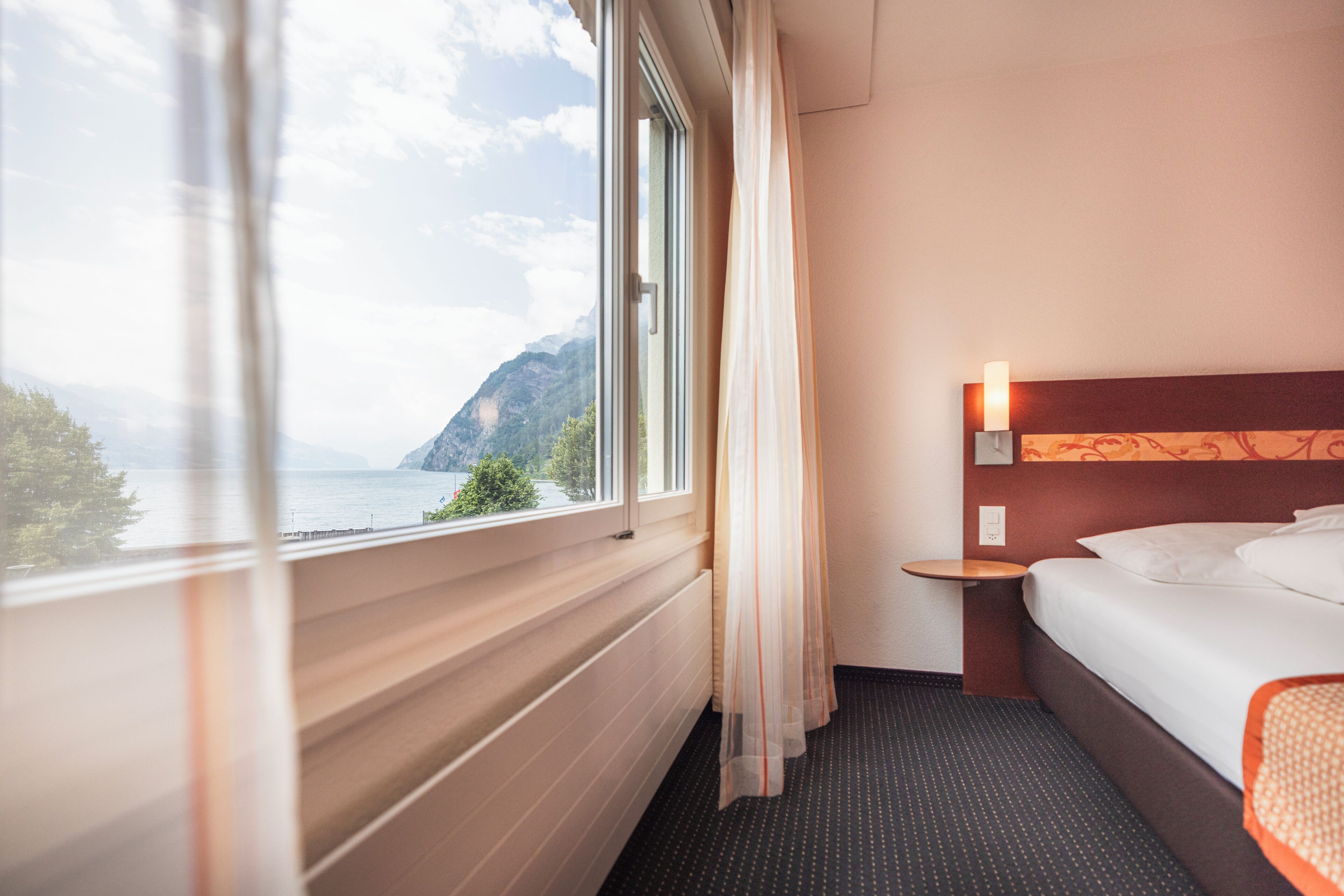 Quarto de hotel com uma janela grande, uma cama, uma mesa de cabeceira e uma luminária fixada na parede.