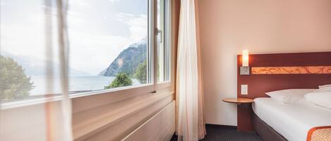 Quarto de hotel com uma janela grande, uma cama, uma mesa de cabeceira e uma luminária fixada na parede.