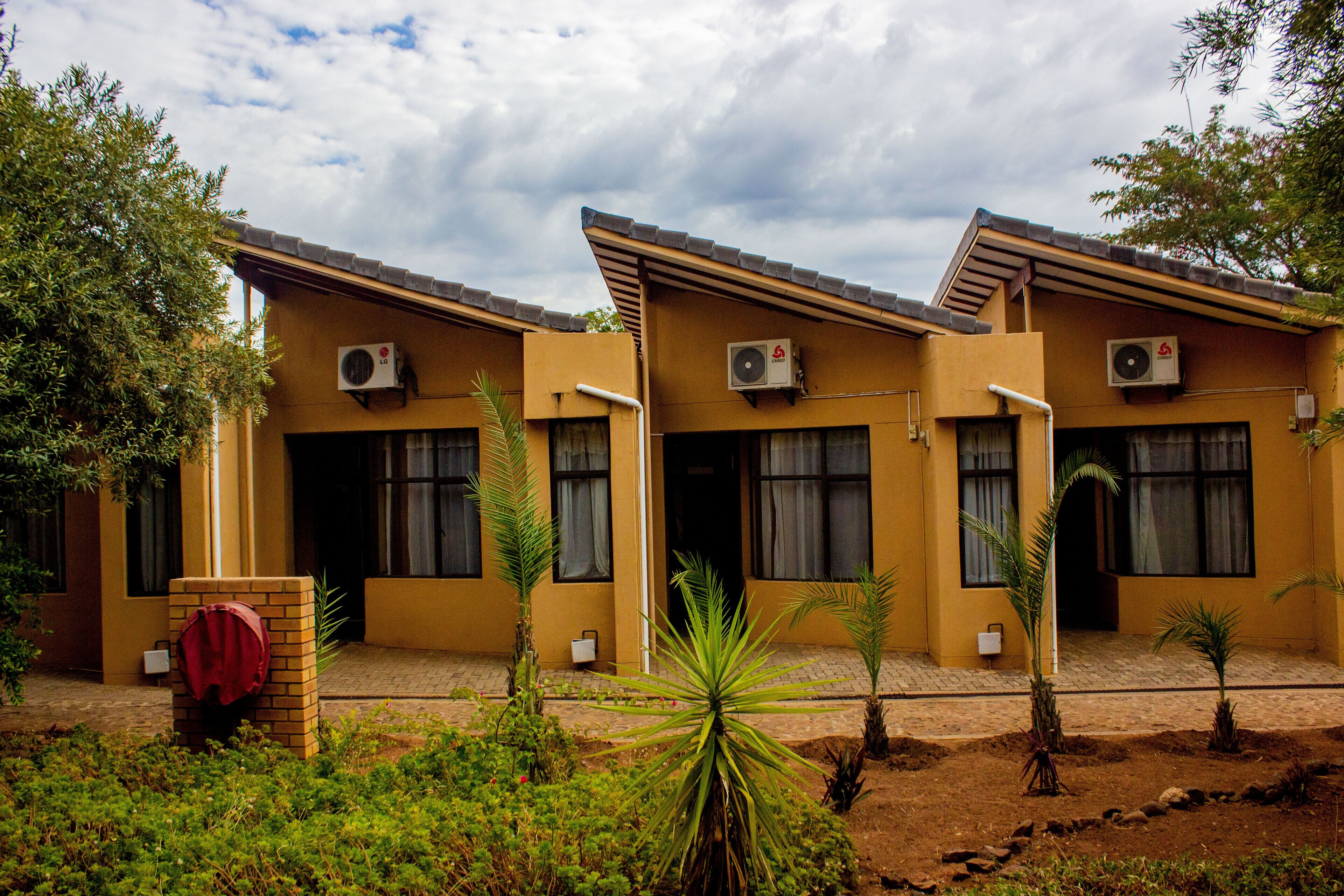 A row of yellow buildings with air conditioning units, a brick entrance, and small palm trees in the foreground.