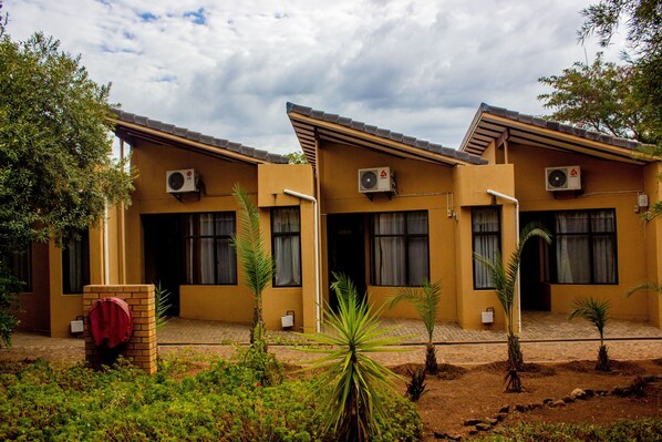 A row of yellow buildings with air conditioning units, a brick entrance, and small palm trees in the foreground. - Wild View Resort (Kasane)