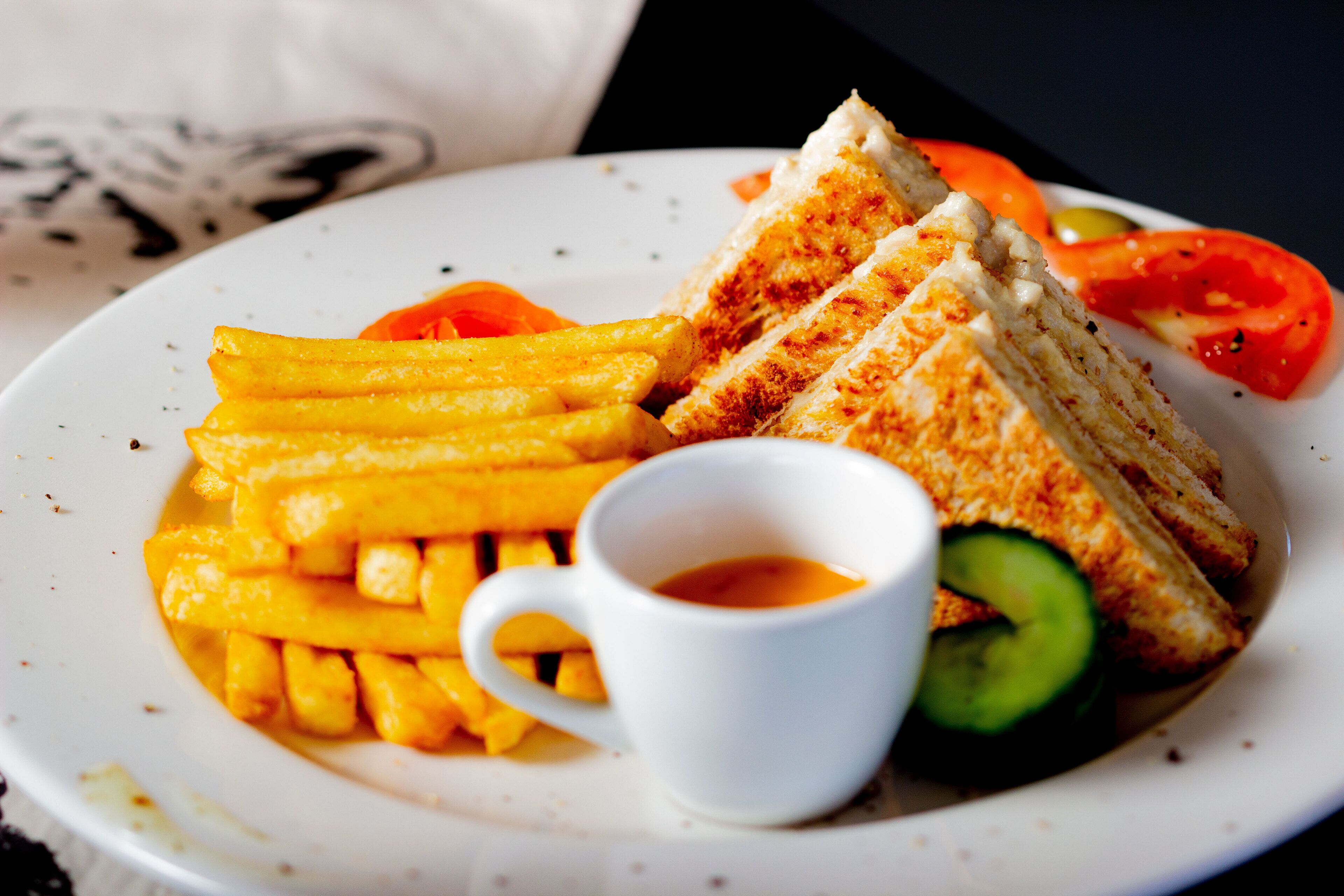 A plate with grilled cheese sandwich, French fries, a slice of tomato, a cucumber, and a cup of sauce.