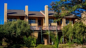 Un hotel de varios pisos con balcones, rodeado de vegetación y un cielo azul despejado.
