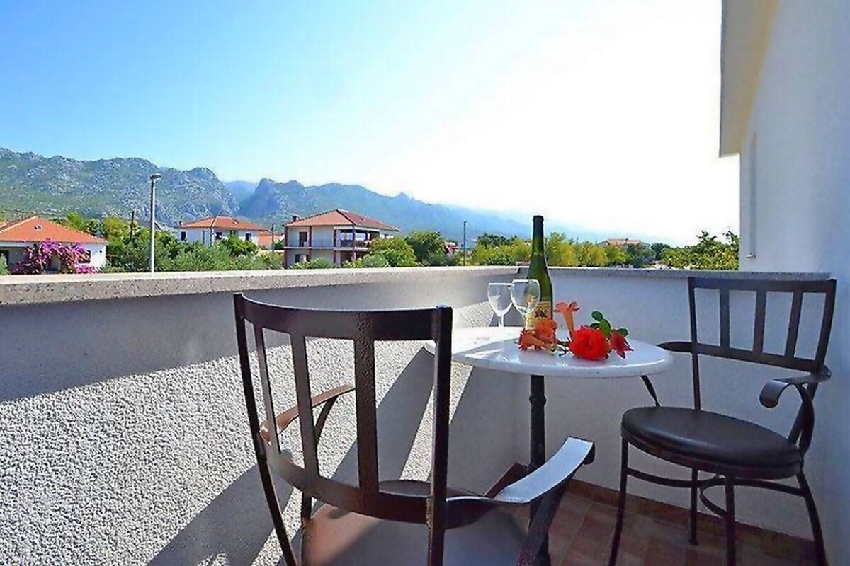 A terrace with a table and chairs, a bottle of wine, and a view of houses and mountains.