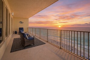 A balcony with a view of the ocean at sunset, a wicker chair, and a patterned rug.