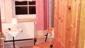 Ein kleiner Raum mit einem Holzschrank, einem Tisch mit Lampe, einem Stuhl und einem Fenster mit Vorhang.