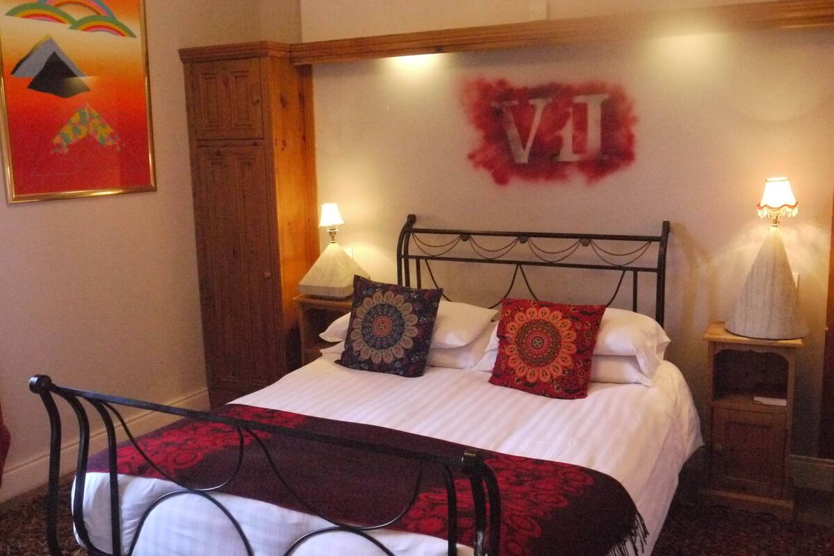 Un lit bien fait, avec un cadre en métal, deux lampes de chevet et une décoration murale représentant la lettre « V » et un cœur rouge.