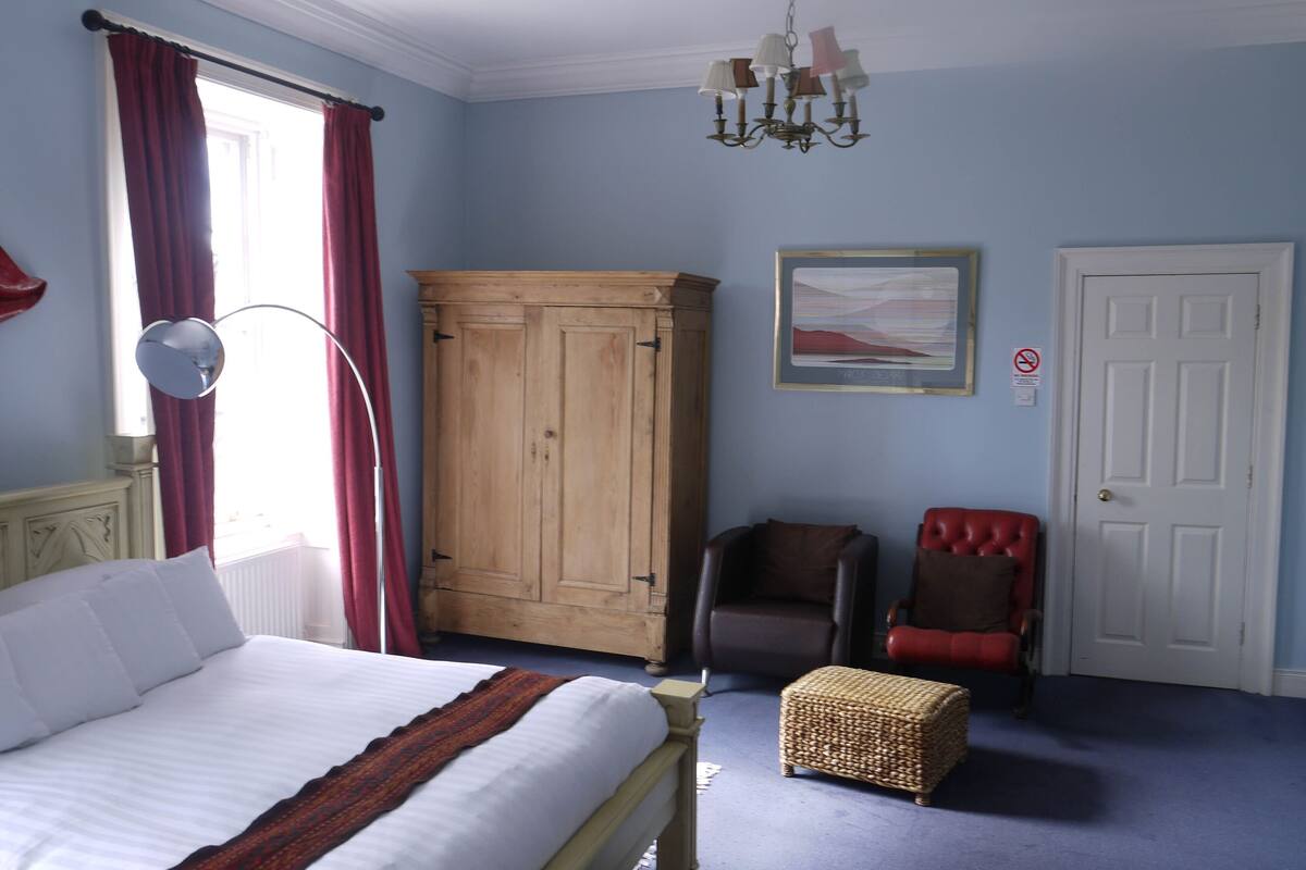 Une chambre à coucher avec un lit, une armoire en bois, un fauteuil rouge, un pouf en osier et un lustre.