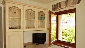 A room with a television on a marble countertop, a cabinet with glass doors, and a window with a view of greenery. - KALIPE' DOLOMITI Appartment (LONGARONE (BL))