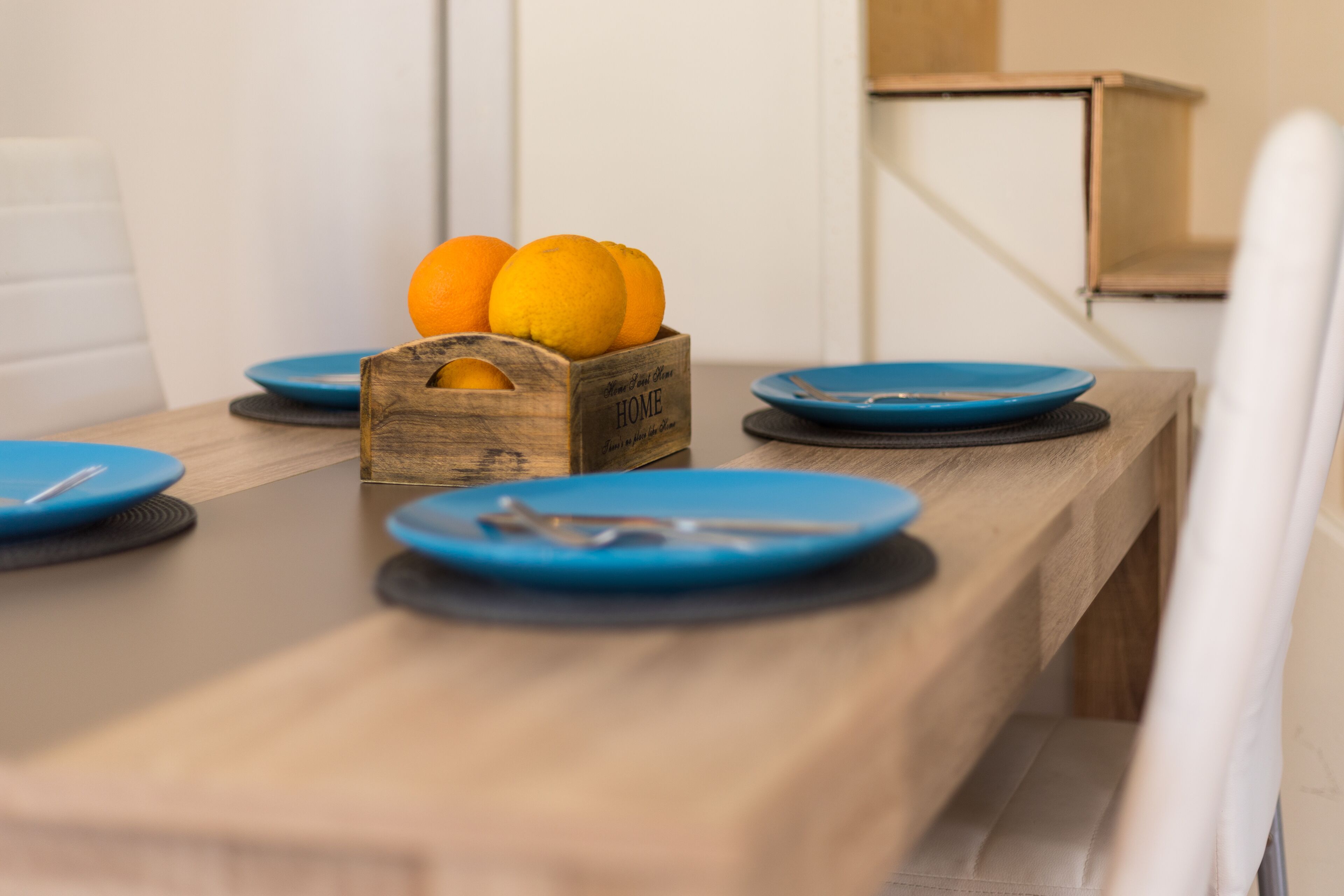 Una mesa de comedor de madera con platos azules y cubiertos, una cesta de frutas de madera con naranjas, y una silla blanca.
