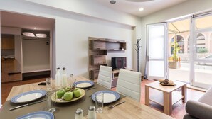 Un comedor moderno con una mesa de madera preparada para cuatro personas, un televisor y vista de la calle a través de puertas de vidrio.