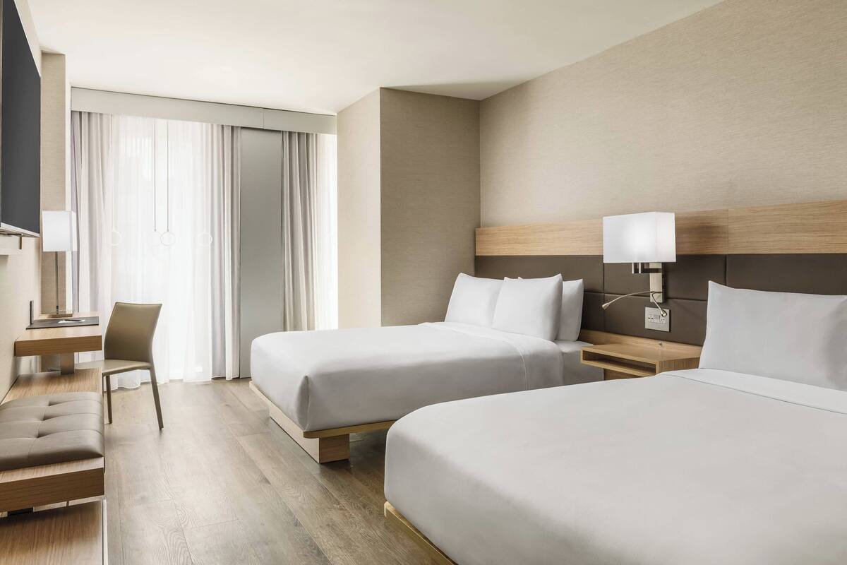 A modern hotel room with two beds, a desk, a chair, and a large window with curtains.