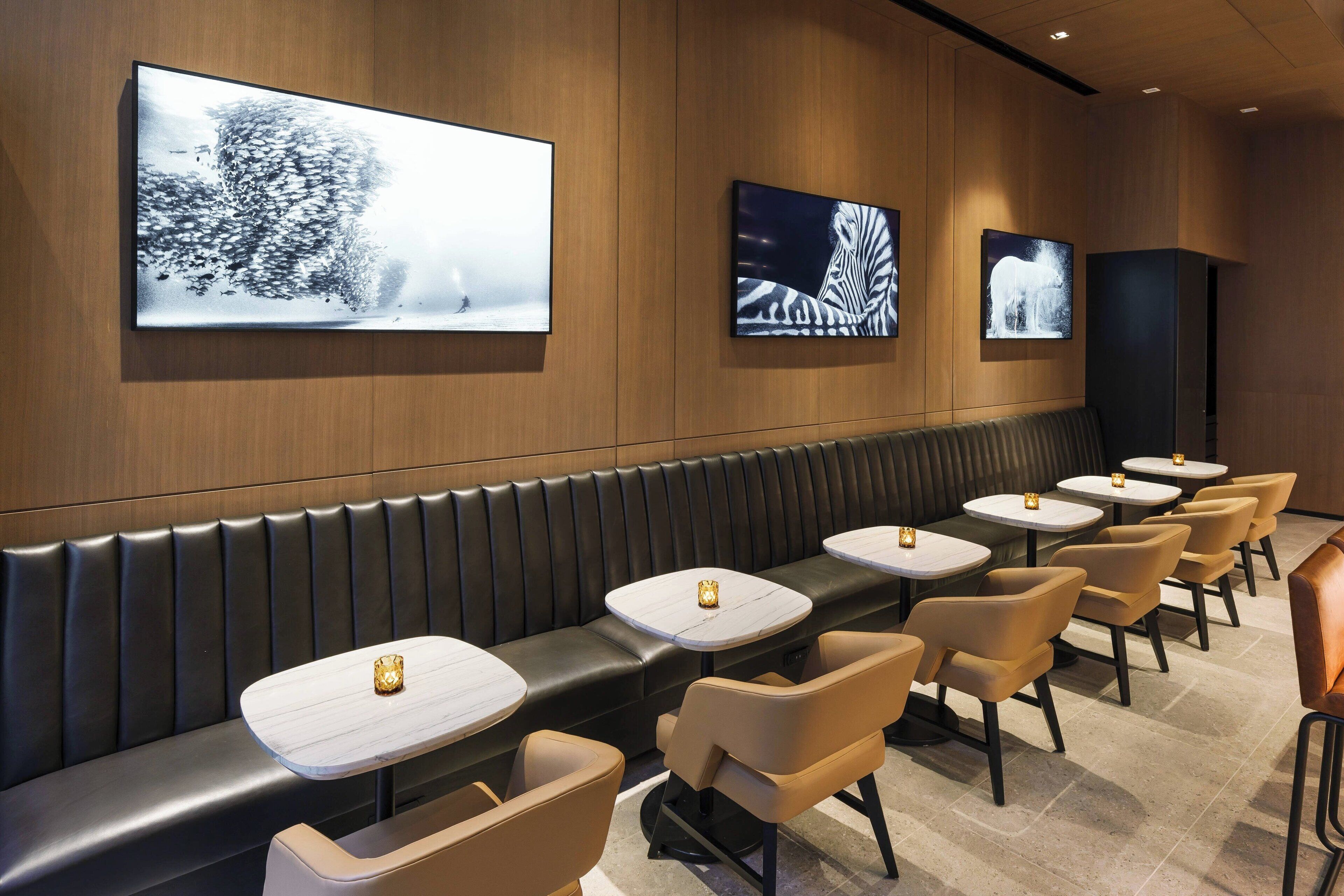 A modern restaurant with a long black leather booth, beige chairs, and small round tables.