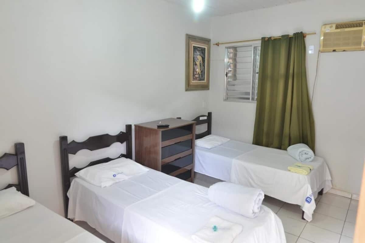 A hotel room with two single beds, a wooden dresser, a framed picture on the wall, and a window with green curtains.