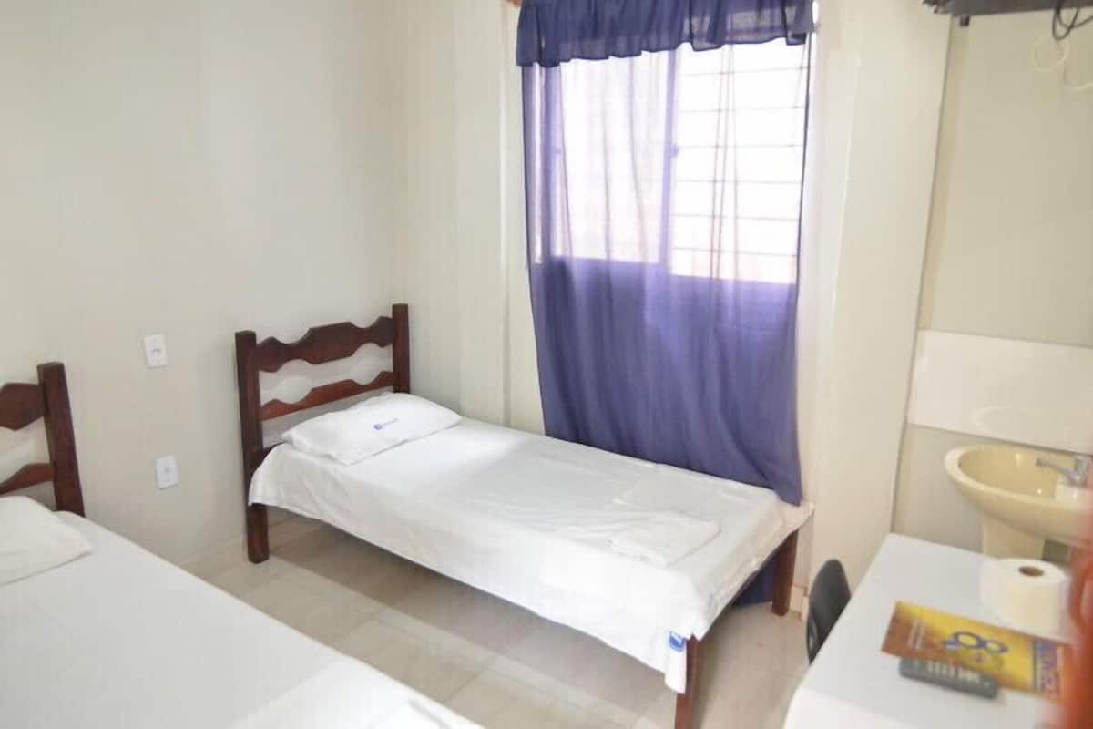 A small, clean room with a single bed, a bedside table, a window with curtains, and a sink.