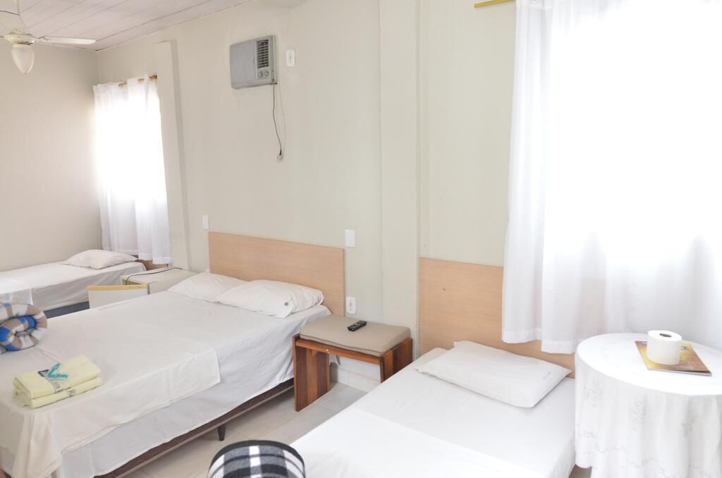 Habitación de hotel con dos camas individuales, una mesita de noche pequeña con un control remoto, aire acondicionado instalado en la pared y una ventana con cortinas.