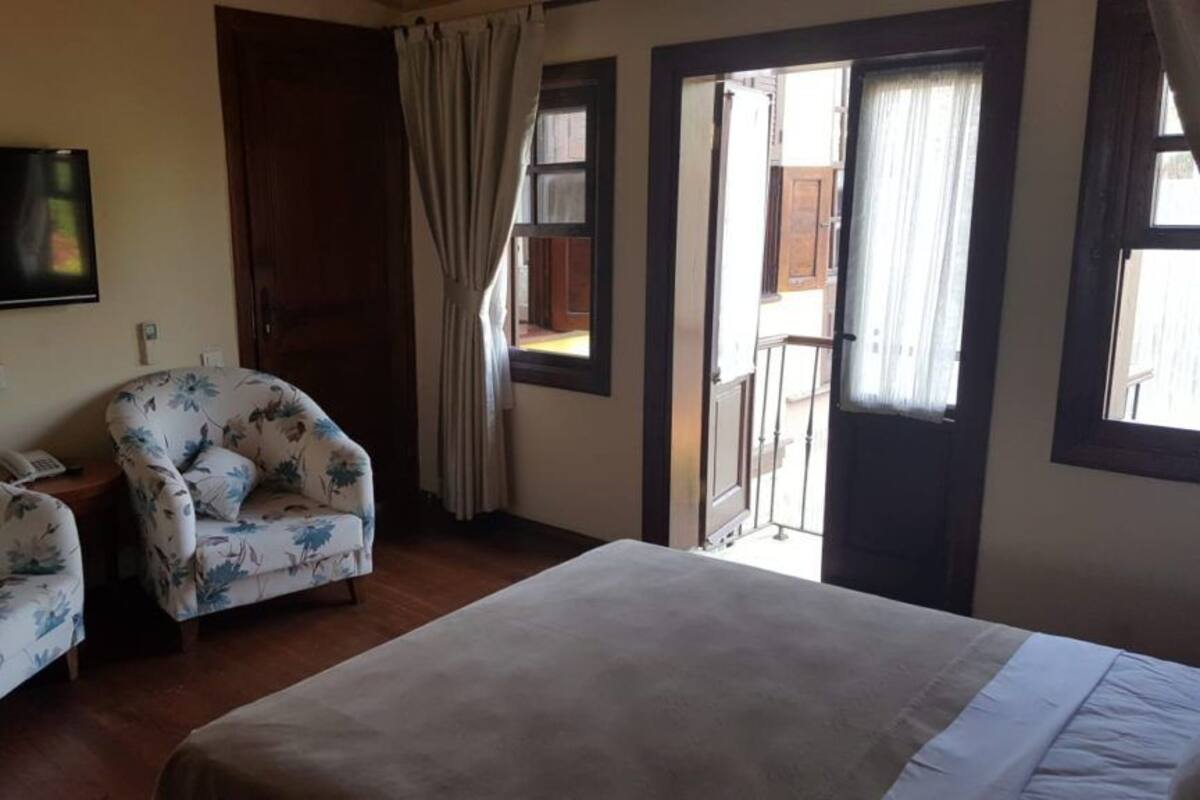 Quarto de hotel com cama, duas poltronas florais, televisão e varanda com cortinas.