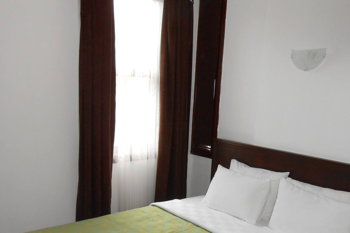 Uma cama de solteiro com colcha verde estampada e almofadas brancas em quarto com cabeceira de madeira e janela com cortinas castanhas.