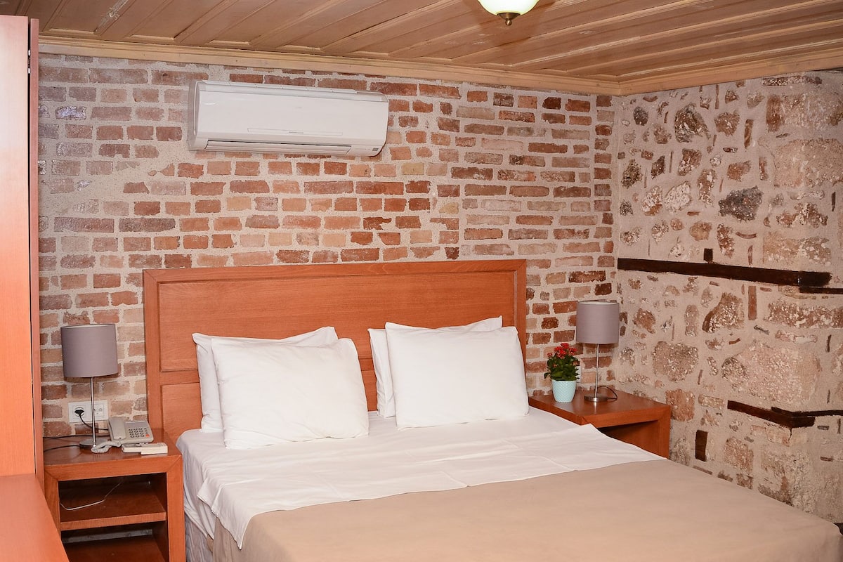 Quarto com cabeceira de madeira, parede com padrão de tijolos, cama branca, duas mesas de cabeceira com luminárias e um aparelho de ar condicionado sobre a cama.