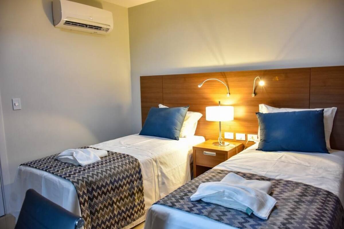 Habitación de hotel con dos camas, cada una con una colcha estampada, una lámpara de noche y un aire acondicionado instalado en la pared.