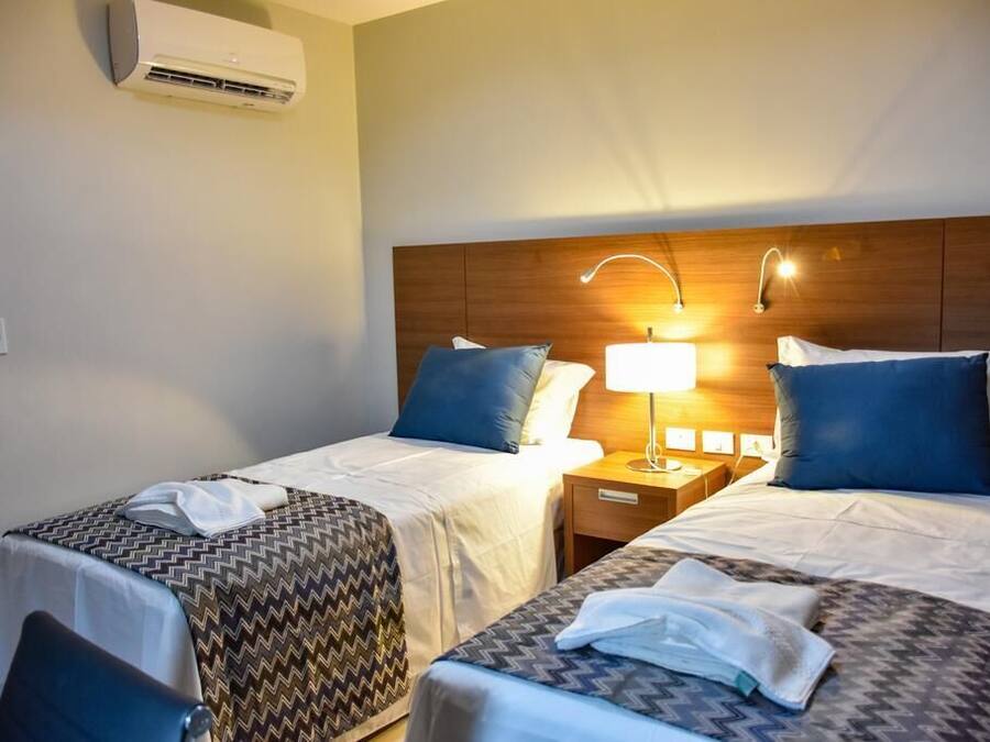 A hotel room with two beds, each with a patterned bedspread, a bedside lamp, and a wall-mounted air conditioner.