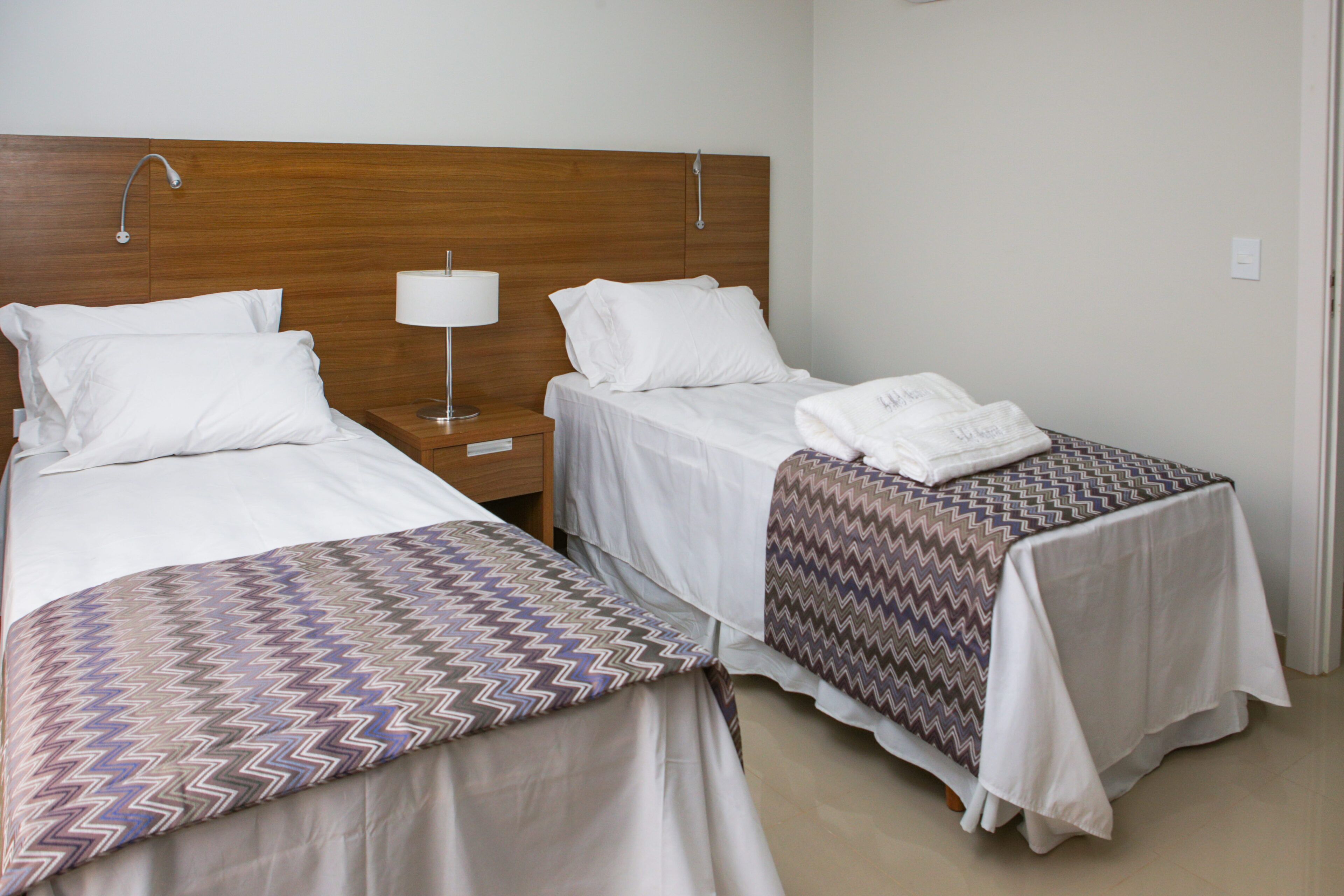 A hotel room with two single beds, a wooden headboard, a nightstand with a lamp, and a bathroom door.