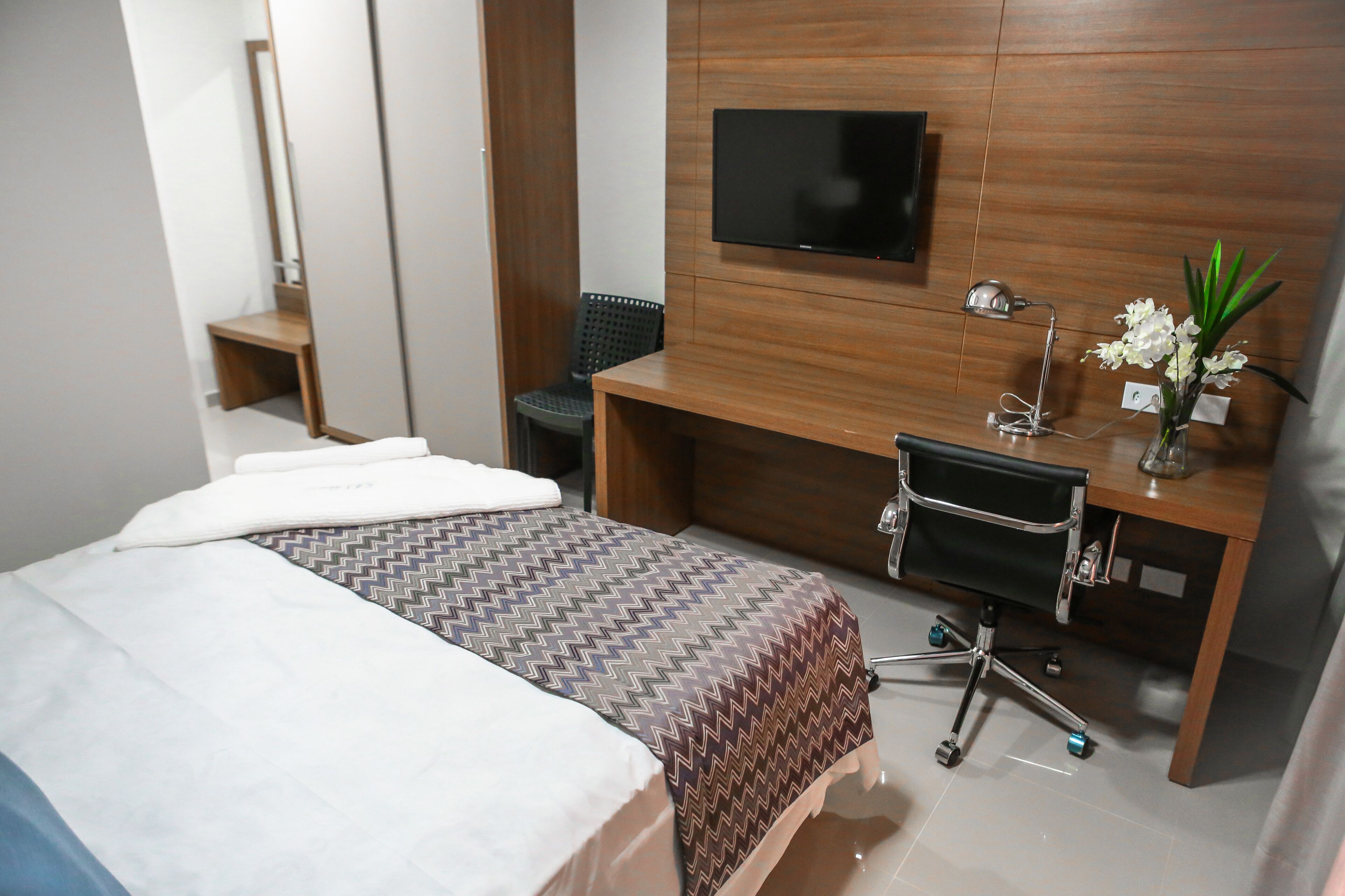 Una habitación de hotel con cama, escritorio y silla, televisor montado en la pared y una pequeña mesa con un jarrón de flores.