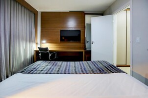 Habitación de hotel con una cama, un escritorio y una silla, un televisor montado en un panel de madera y una puerta que da a otra habitación.