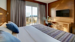 Habitación de hotel con una cama grande, un escritorio, un televisor y vistas de la ciudad a través del ventanal.