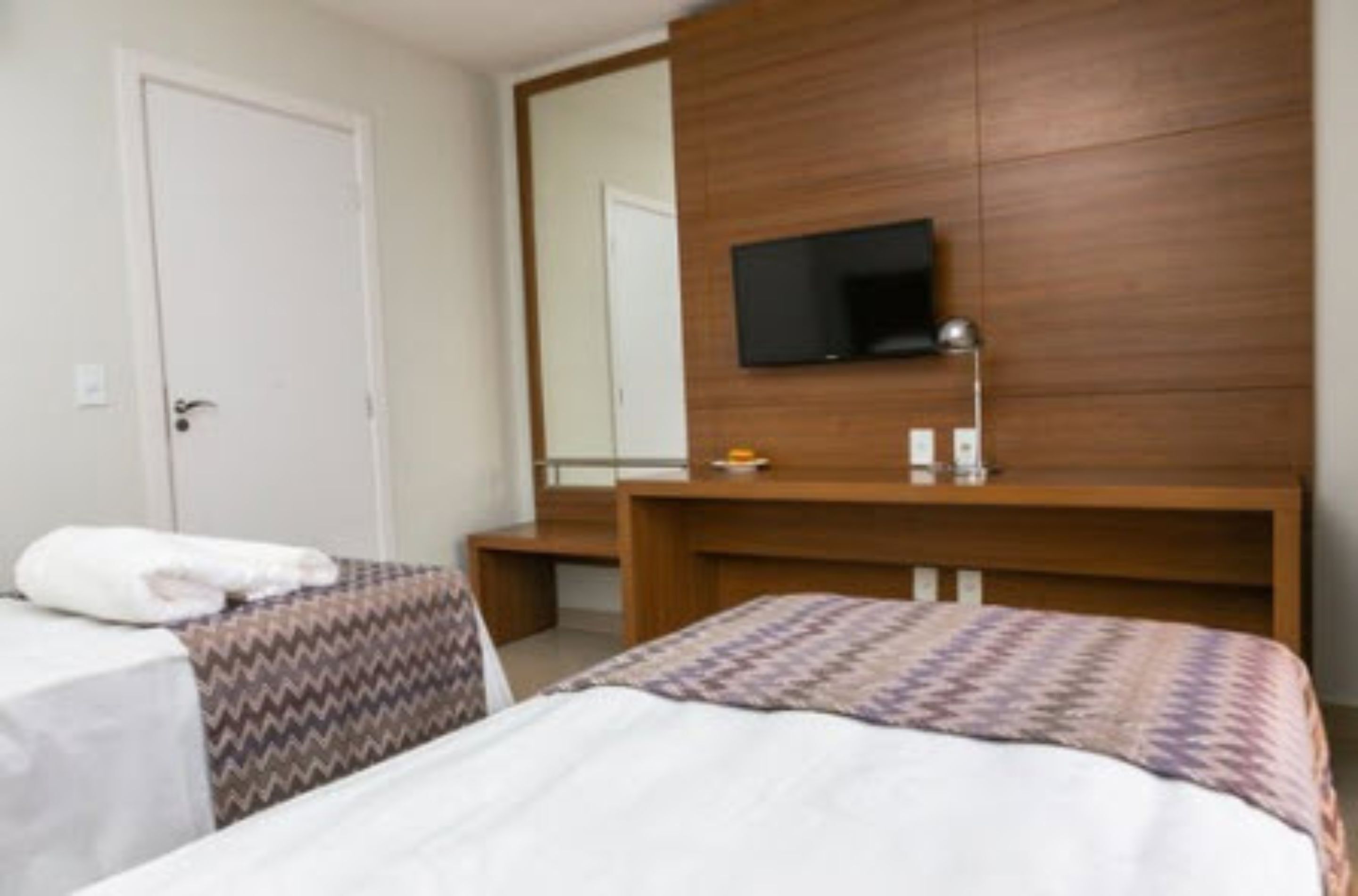 A hotel room with two beds, a wooden headboard, a flat-screen TV mounted above the headboard, and a small table with a lamp and a bottle.