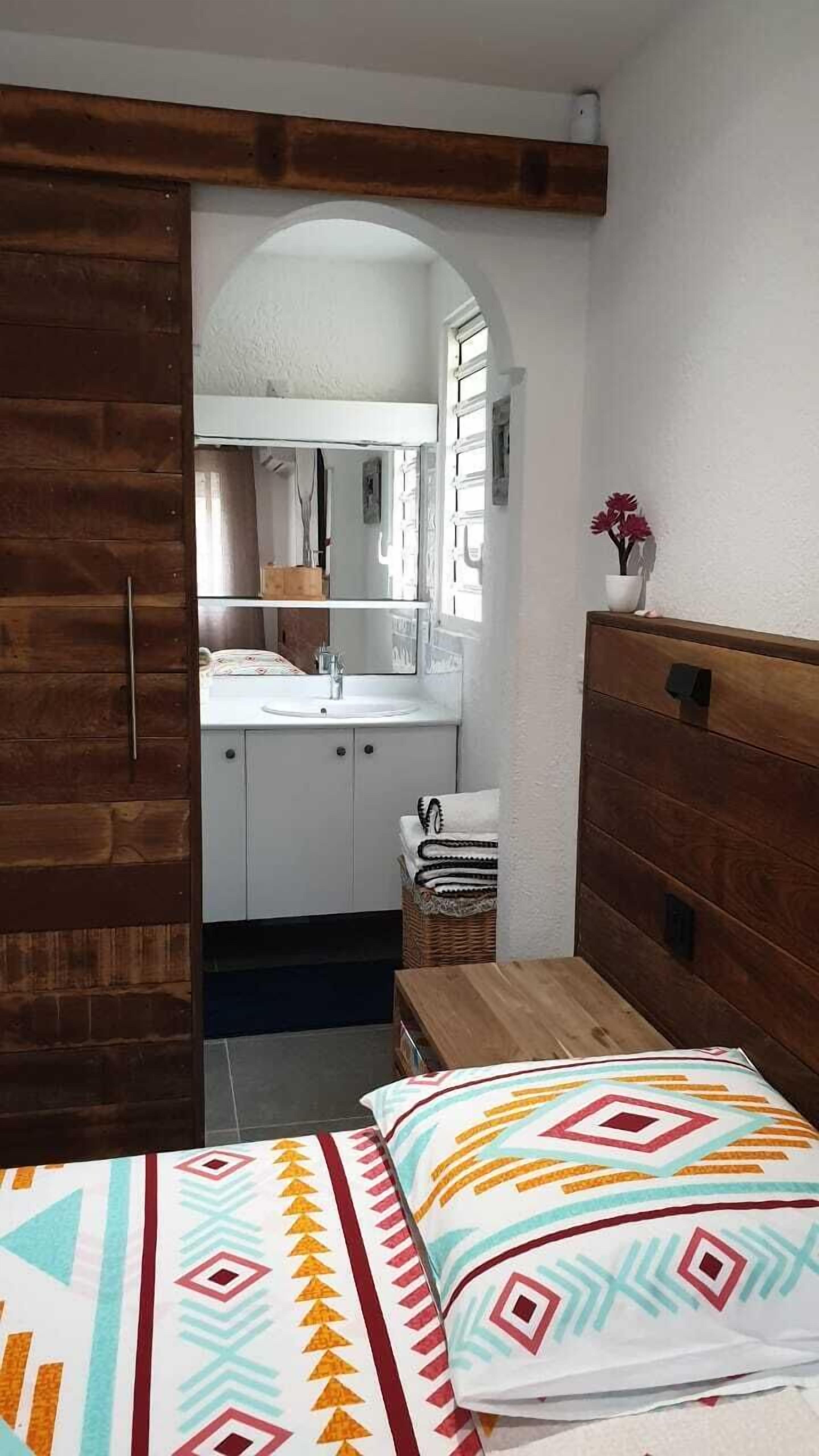 Une chambre avec une armoire en bois, un lit agrémenté d’oreillers à motifs, une petite table de chevet et une vue sur la salle de bain à travers une arche.