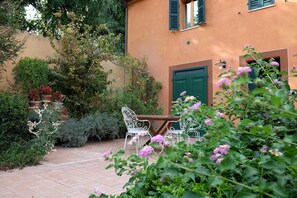 Un patio con una mesa y sillas, rodeado de abundante vegetación y un edificio con puerta verde.