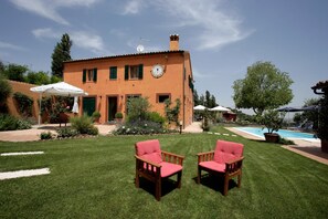 Una villa con piscina, dos sillas rojas y un césped cuidado.