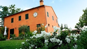 A two-story building with a clock on the front wall, surrounded by greenery and white flowers. - Poggio Montali (Monte Roberto)