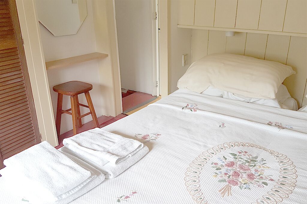 Quarto com uma cama, um banquinho de madeira e uma parede branca.
