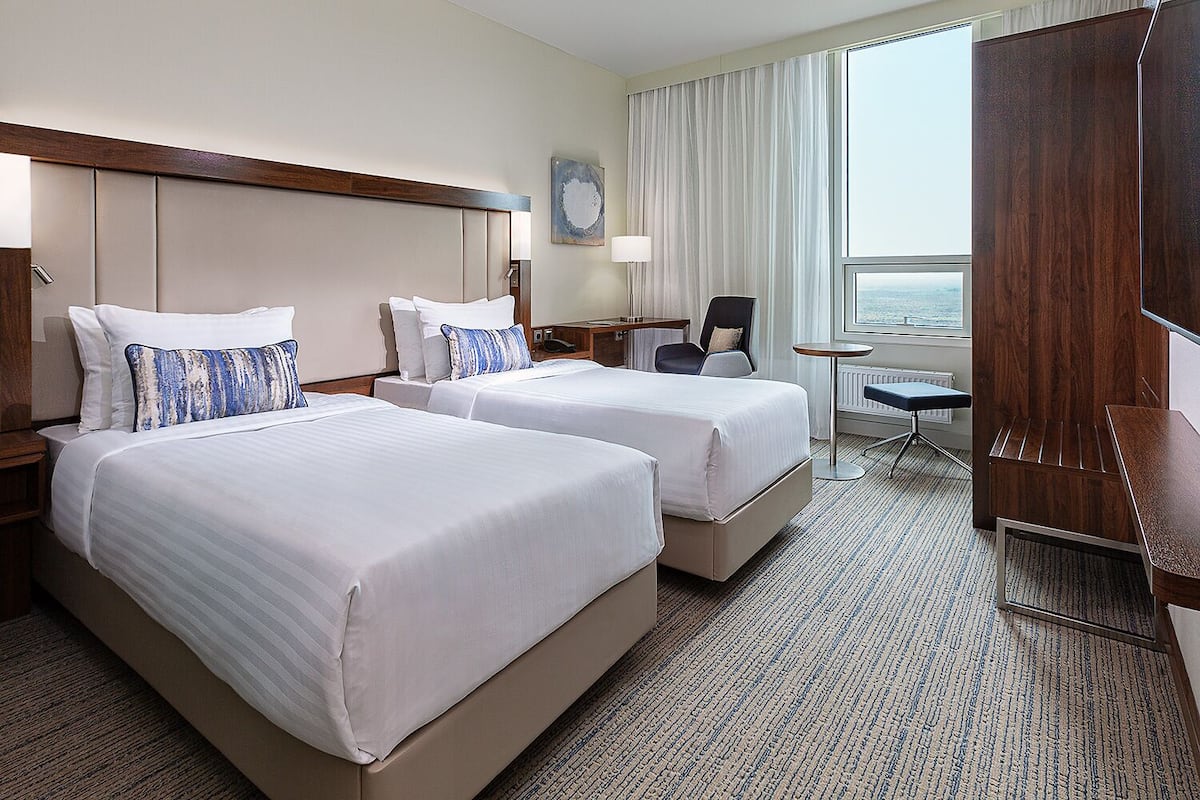 Ein Hotelzimmer mit zwei Betten, einem Schreibtisch, einem Stuhl, einem kleinen Tisch, einem Fernseher und einem großen Fenster.