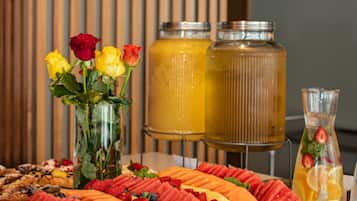 Une table de buffet garnie d’une variété de fruits, un vase rempli de fleurs et deux grands distributeurs de boissons.