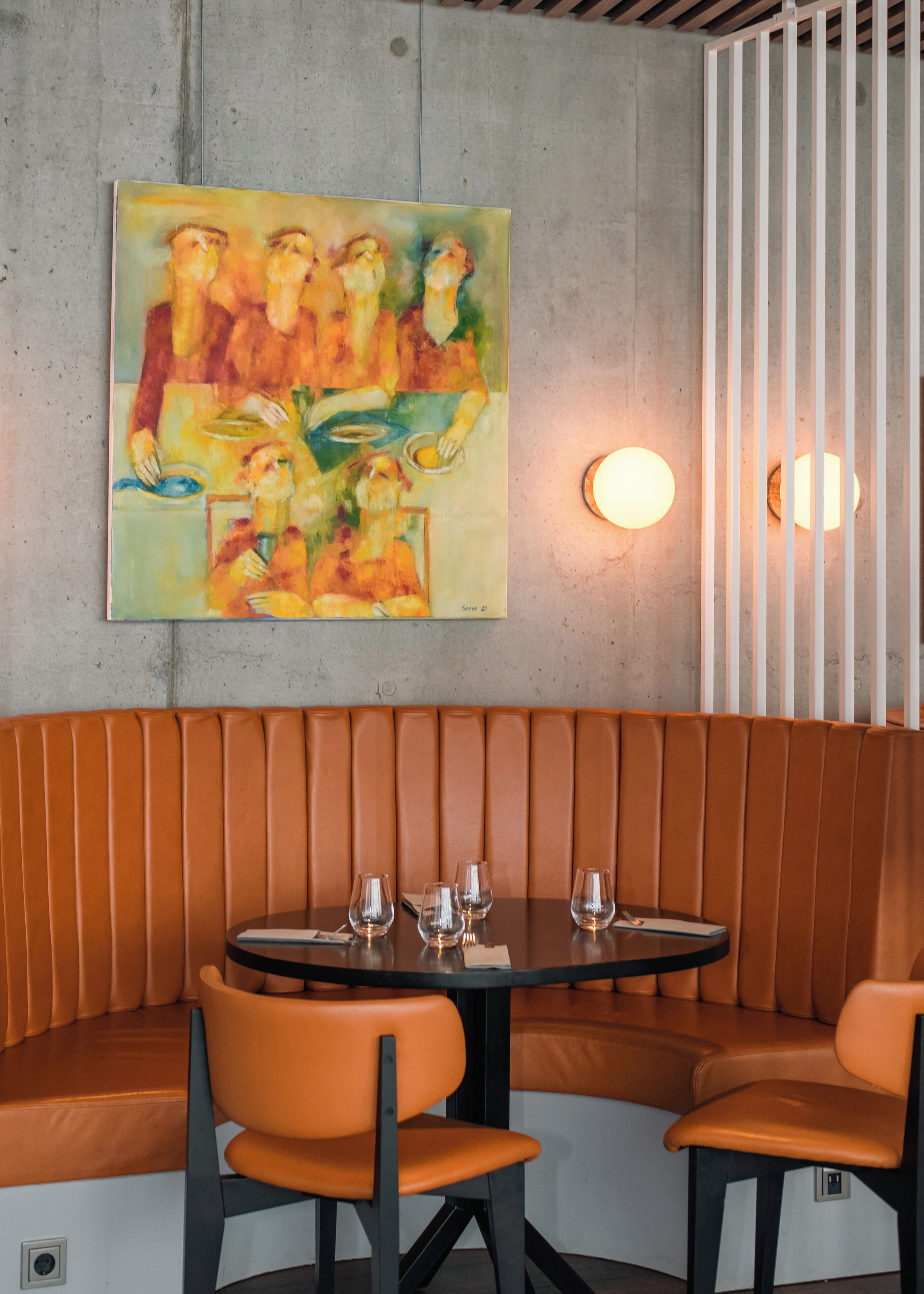 Interior de un restaurante con una zona de asientos tipo cabina, una mesa redonda con vasos y cubiertos, y un cuadro en la pared.