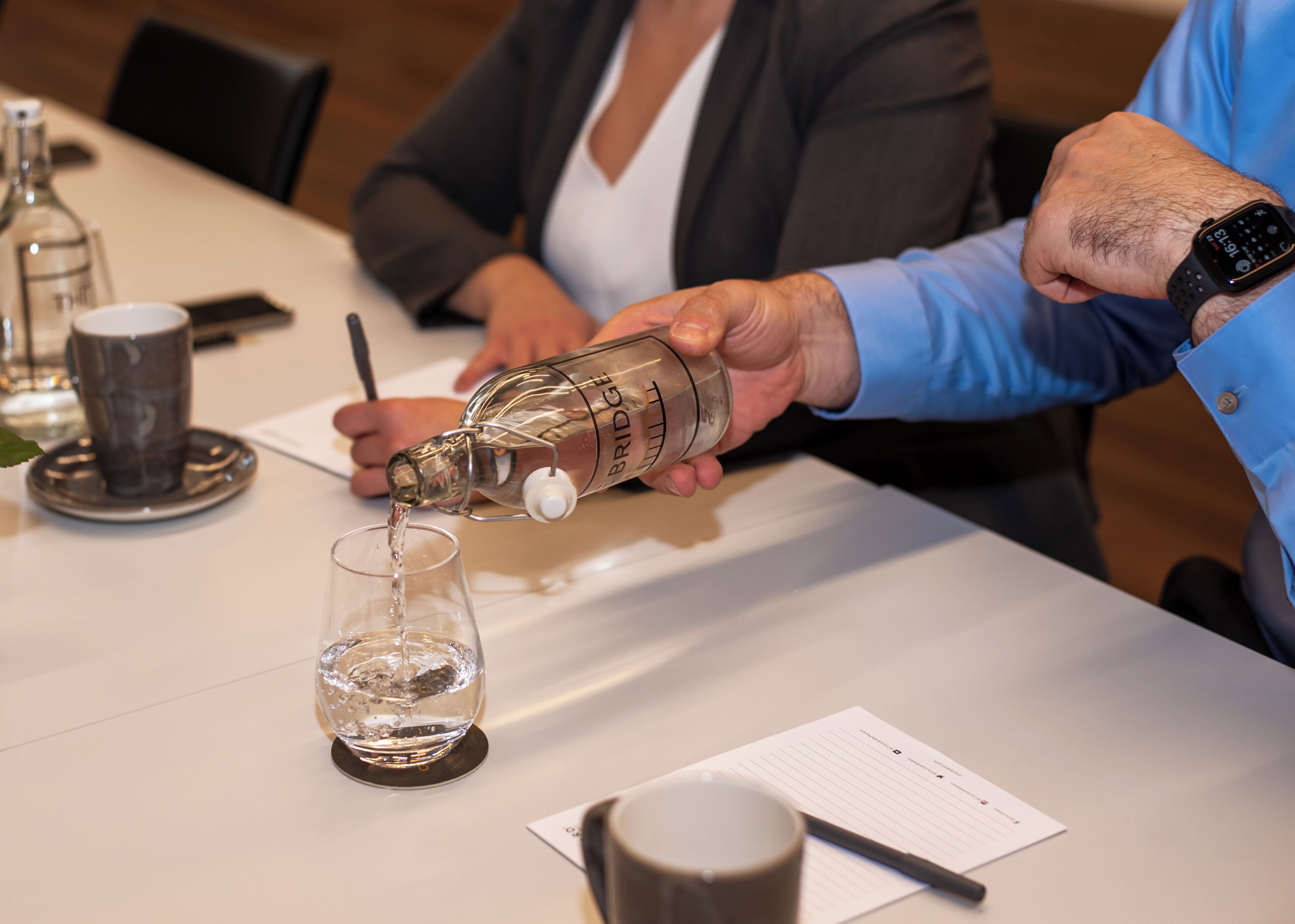 Une personne verse un liquide transparent d’une bouteille dans un verre posé sur une table.