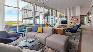 Un moderno vestíbulo de hotel con ventanas amplias, una zona de estar con sofás y sillones, una mesa de centro y una recepción.